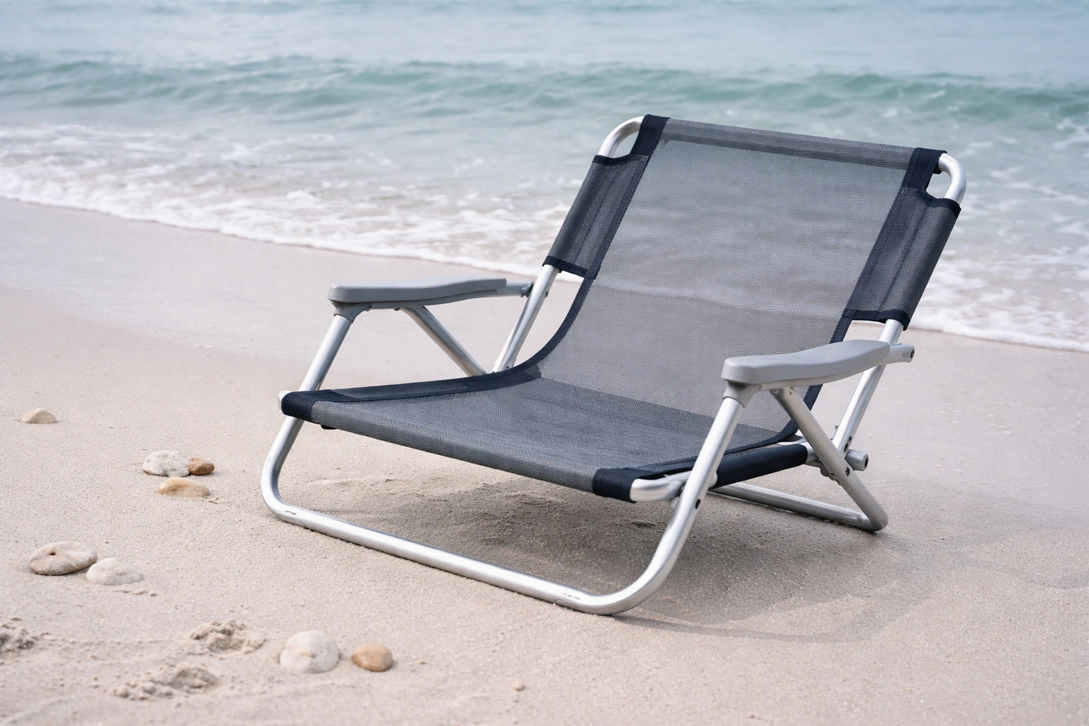 Low-slung beach chair with aluminum frame and mesh seat on white sand at the waterline with gentle waves