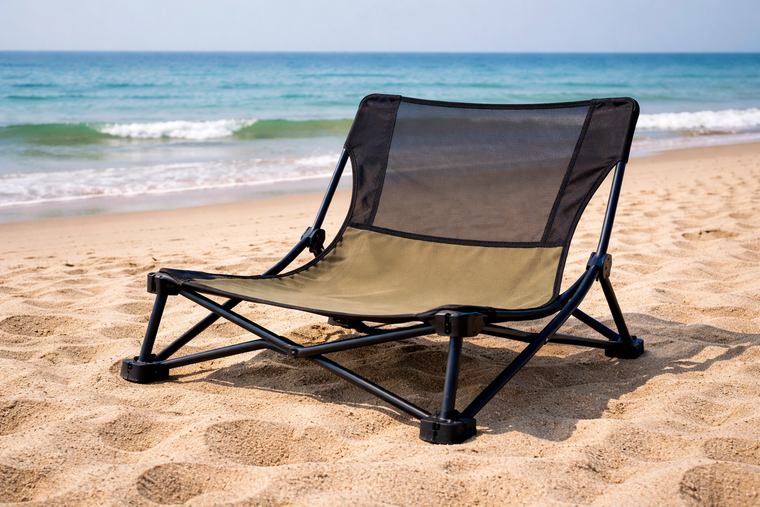 Low-profile steel-frame camping chair with mesh back panel on sandy beach near waterline with ocean waves and clear sky