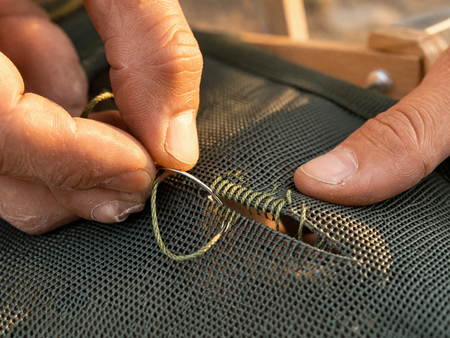 Close-up of hands whipstitching a torn mesh seam on camping chair with curved needle and green thread