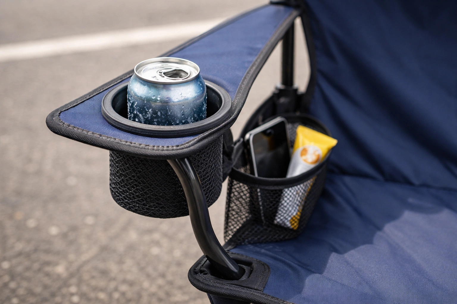 Chair armrest showing insulated cup holder with cold beverage condensation and mesh pocket with phone and snacks on asphalt
